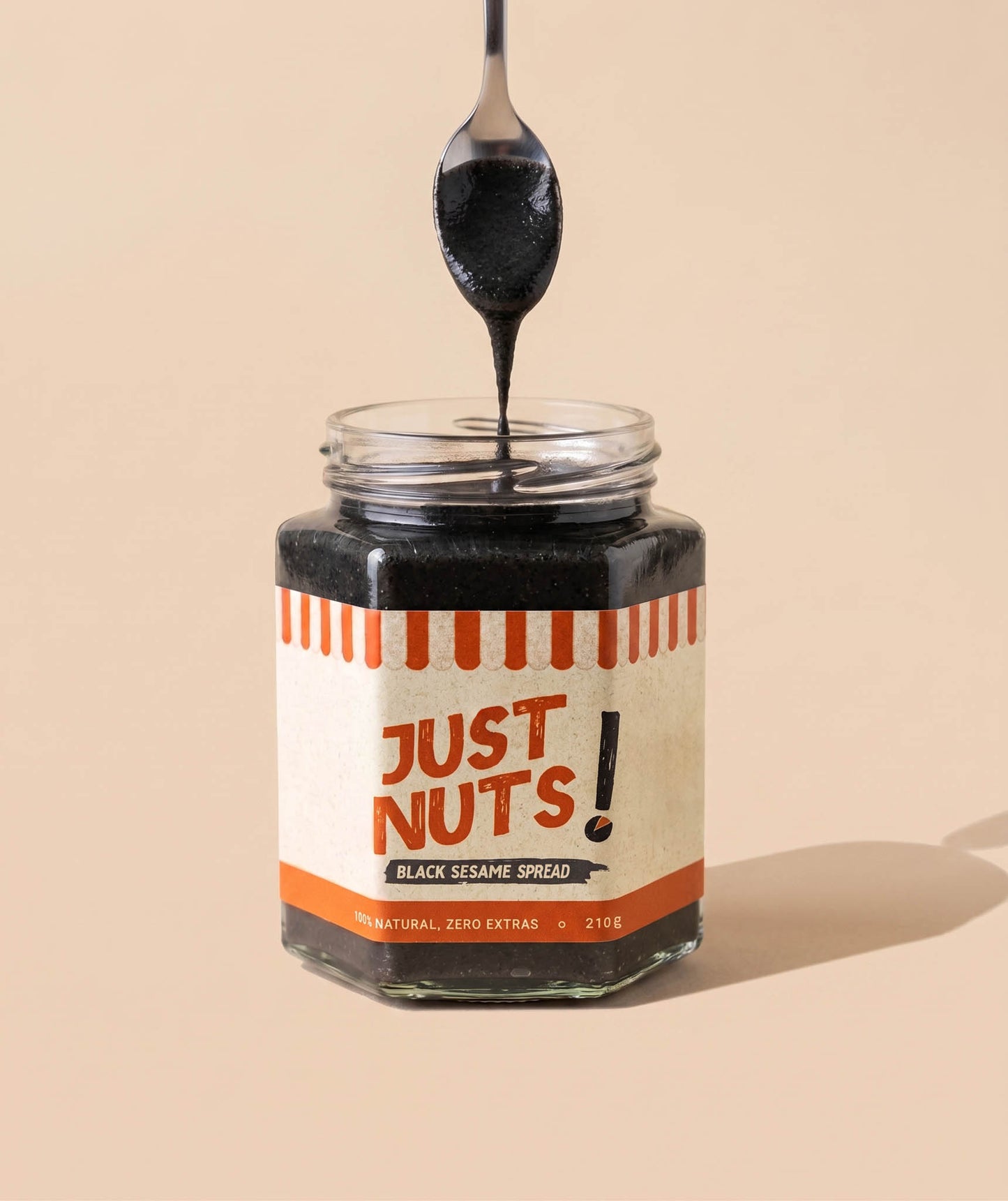 A jar of black sesame spread with a spoon lifting the thick dark spread above the open jar, set against a solid beige background. The label reads 'JUST NUTS! BLACK SESAME SPREAD'.