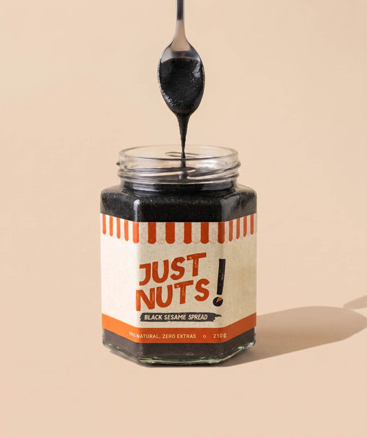 A jar of black sesame spread with a spoon lifting the thick dark spread above the open jar, set against a solid beige background. The label reads 'JUST NUTS! BLACK SESAME SPREAD'.