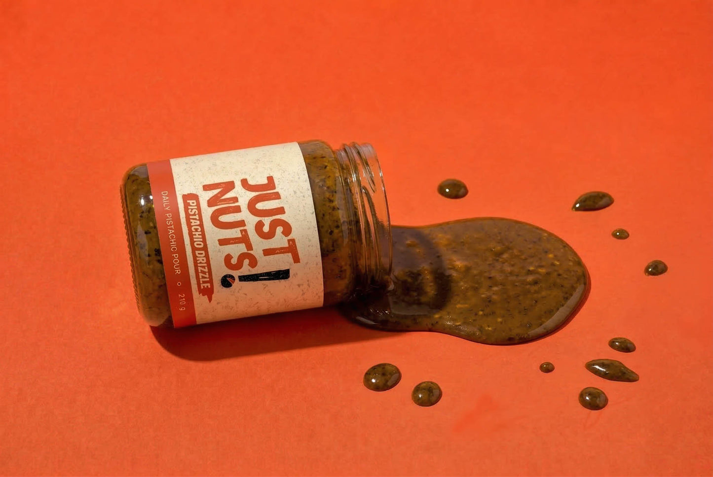 A clear glass jar of Just Nuts Spreading Dough lies on its side with dark brown spread spilled out onto a solid orange-red background. The white label features red text reading 'JUST NUTS'  made in christchurch, best for pistachio matcha latte